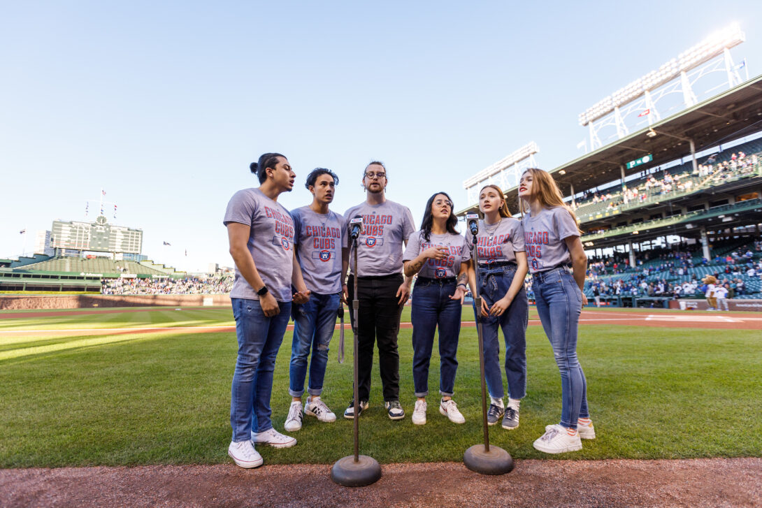 Six UIC students singing the National Anthem in gray Cubs shirts on the infield of Wrigley Field and the iconic scoreboard in the background
